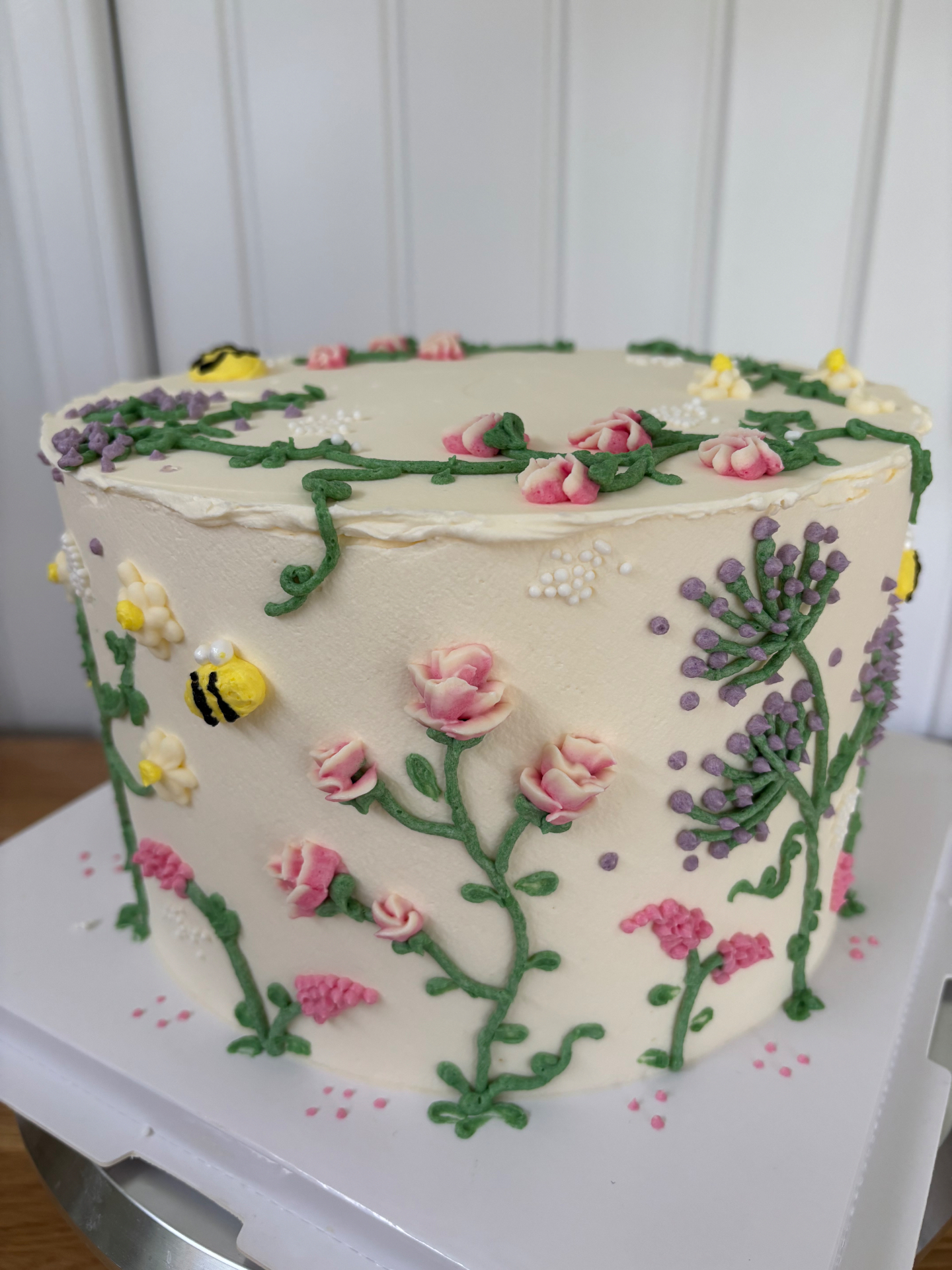 Floral celebration cake with piped pink roses, green vines, lavender sprigs and fondant bees