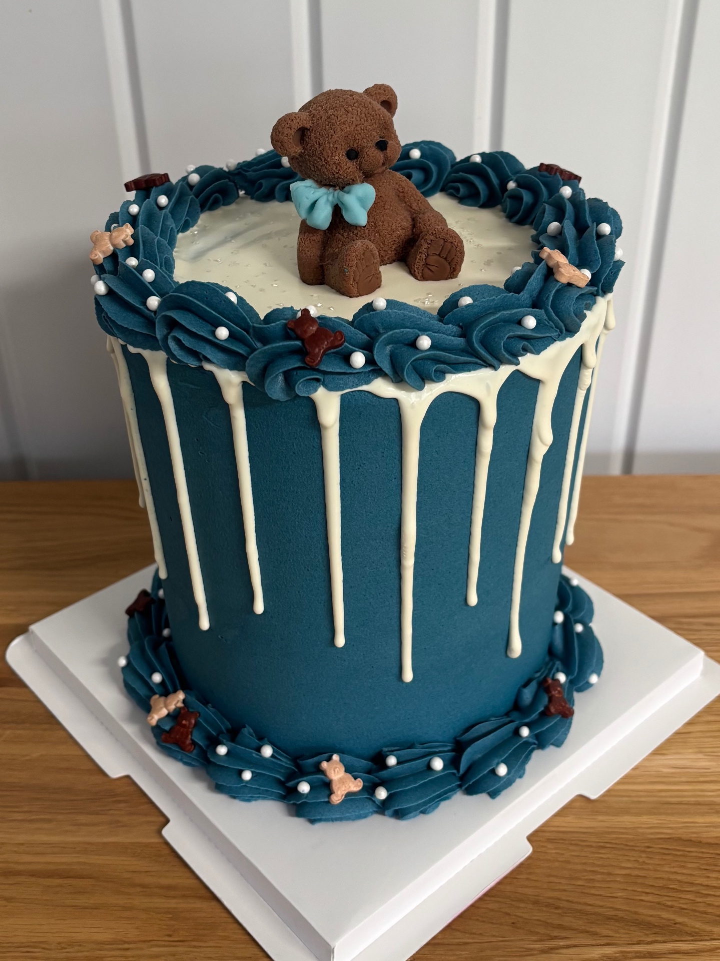 Teal drip cake topped with a sculpted teddy bear wearing a pastel blue bow tie, piped buttercream borders and sugar pearls