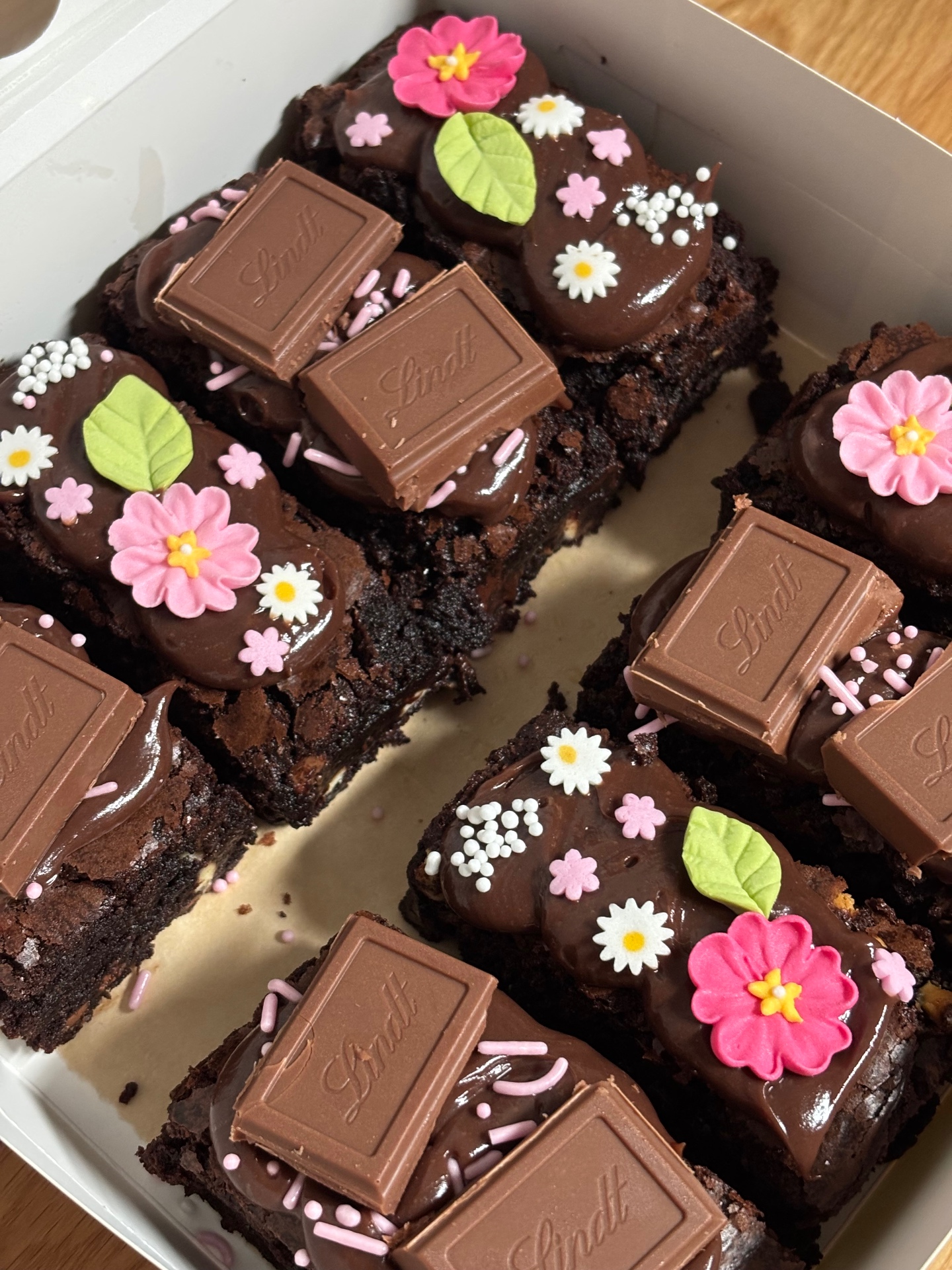 Box of chocolate brownies topped with Lindt squares, pink blossoms, daisies and green leaves